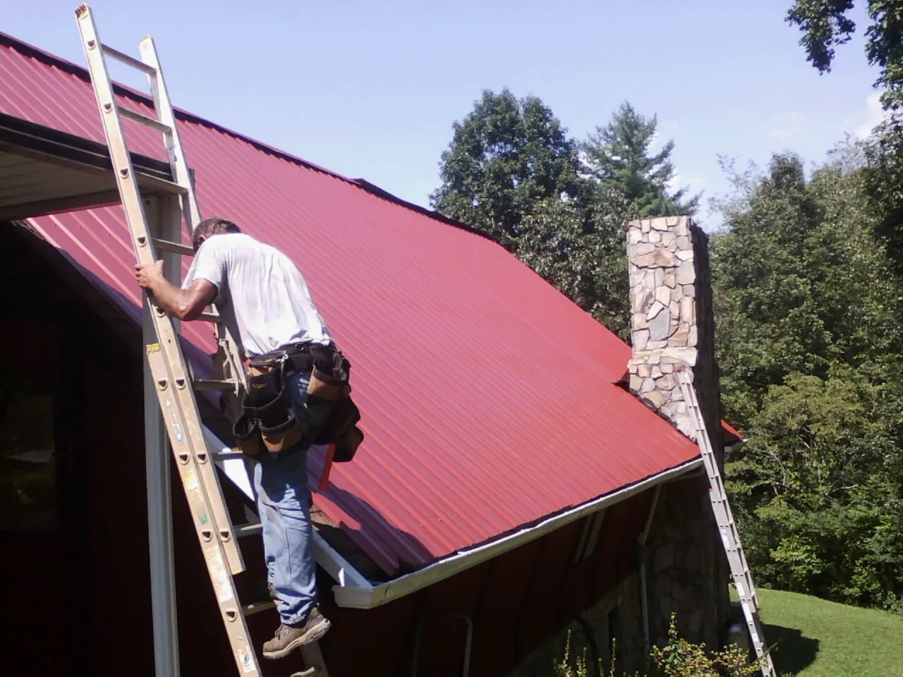 Roofer on a ladder beside a red metal roof and stone chimney — Soffit & Fascia Repair by Roofing Brothers in Homedale
